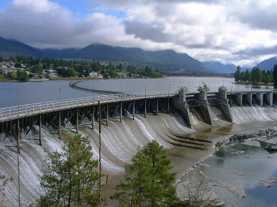 [The dam is very wide across, but is relatively short in height. There are a number of visible poles supporting the upper deck of the dam and thin streams of water pass around them across the concrete. There are plenty of evergreens and buildings behind the dam and a mountain range in the distance behind it.]